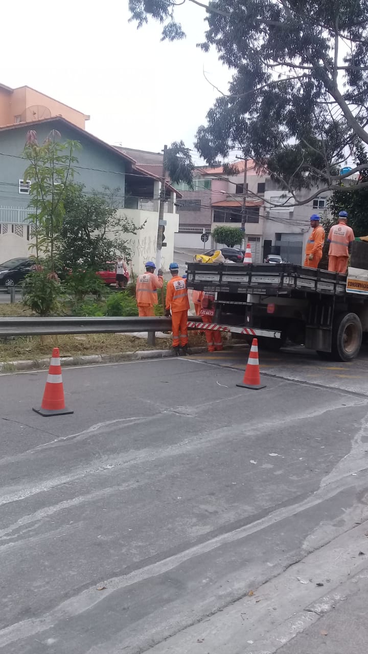 Dois cones dispostos na rua cercam o caminhão da Prefeitura. Há 3 funcionários na calçada e 2 em cima do caminhão, todos de uniforme laranja e capacete azul.